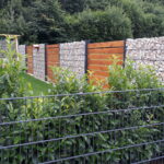 Ein trister Garten mit kurzem Rasen, fehlenden Blüten, giftigem Kirsch-Lorbeer im Vordergrund, Gabionen-Wand. Im Hintergrund sieht man einen mit Kunststoffplane "geschmückten" Zaun, auf die die Pflanzen nur noch aufgedruckt sind.  Pflegeleicht - aber dieser Garten hat dem Klimawandel nichts entgegen zu setzen.